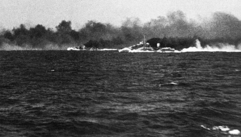Moments before coming under attack, the British cruiser Gloucester is unable to take advantage of a thick, black smokescreen  laid by the cruisers Ajax and Perth. Other British warships have already been forced to take evasive action as shells from the Italian battleship Vittorio Veneto crash nearby.