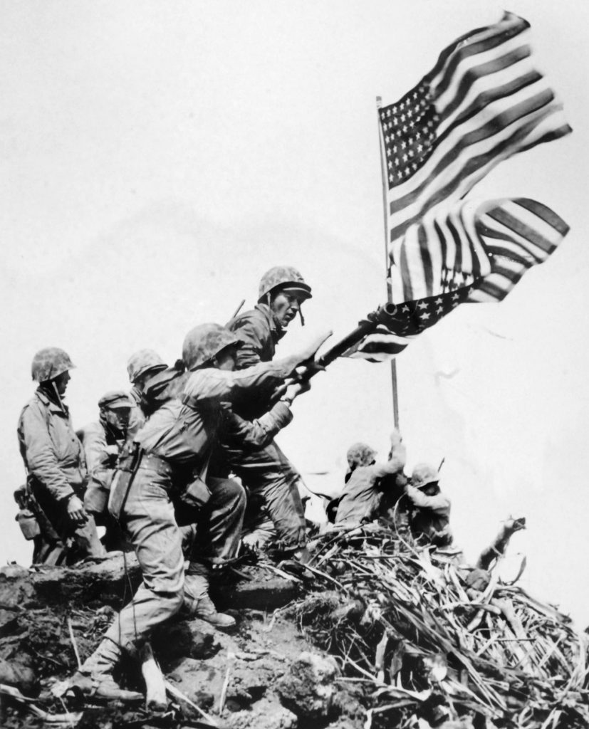 The first, smaller flag (foreground) raised on Iwo Jima’s Mount Suribachi has been replaced by a larger one visible to all the troops on the island. But the battle would go on for nearly three months and cost almost 7,000 Marines their lives. 