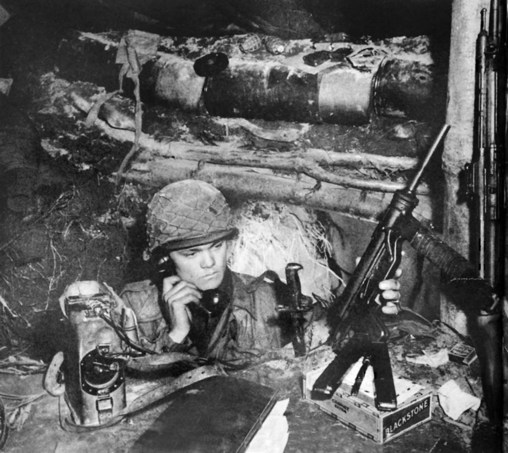 A 42nd Division soldier mans his foxhole while communicating on a field telephone. He holds an M3 submachine gun, nicknamed the grease gun.