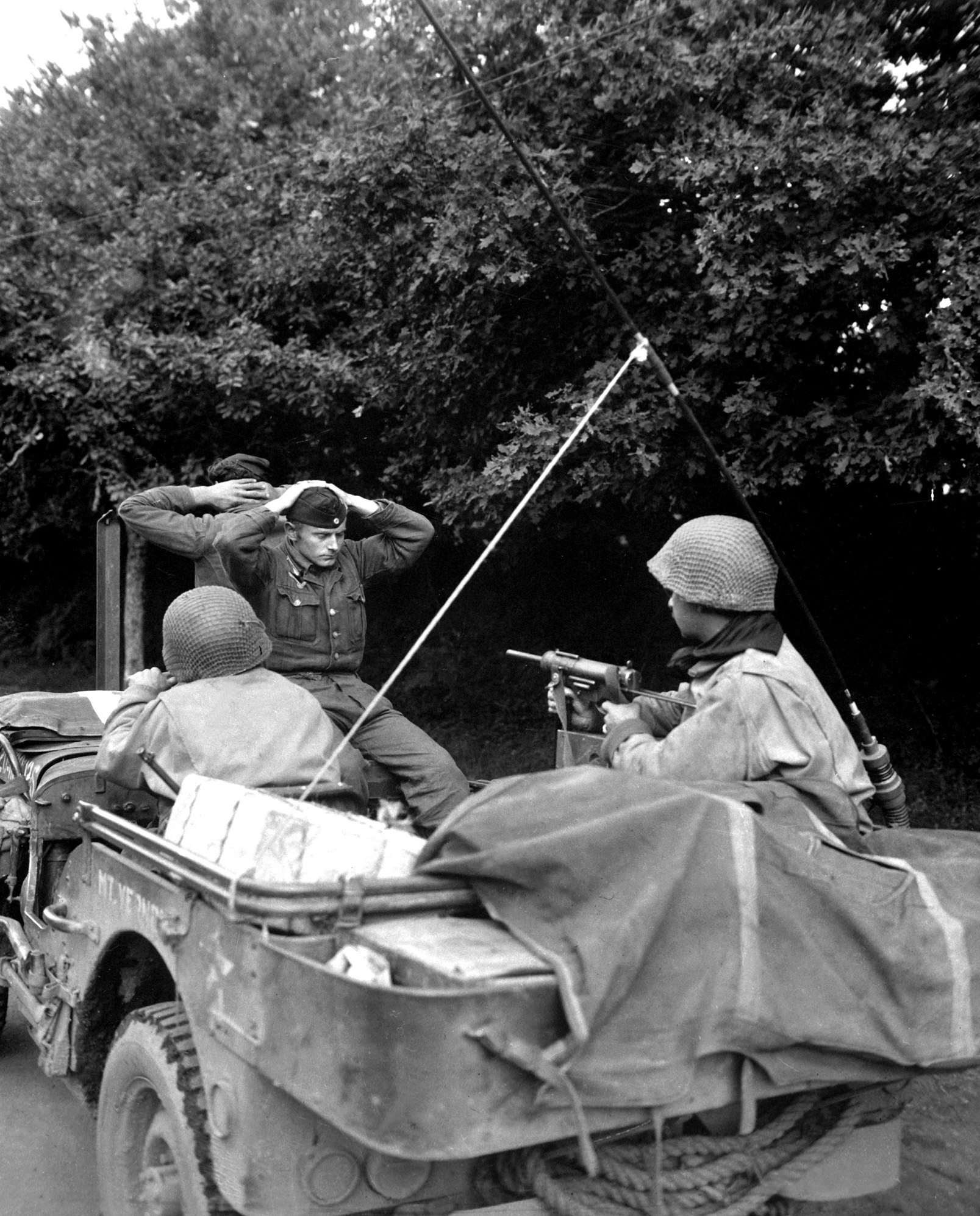 Their hands behind their heads, two German prisoners of war are guarded by an American soldier keeping watch and armed with an M3 Grease Gun.