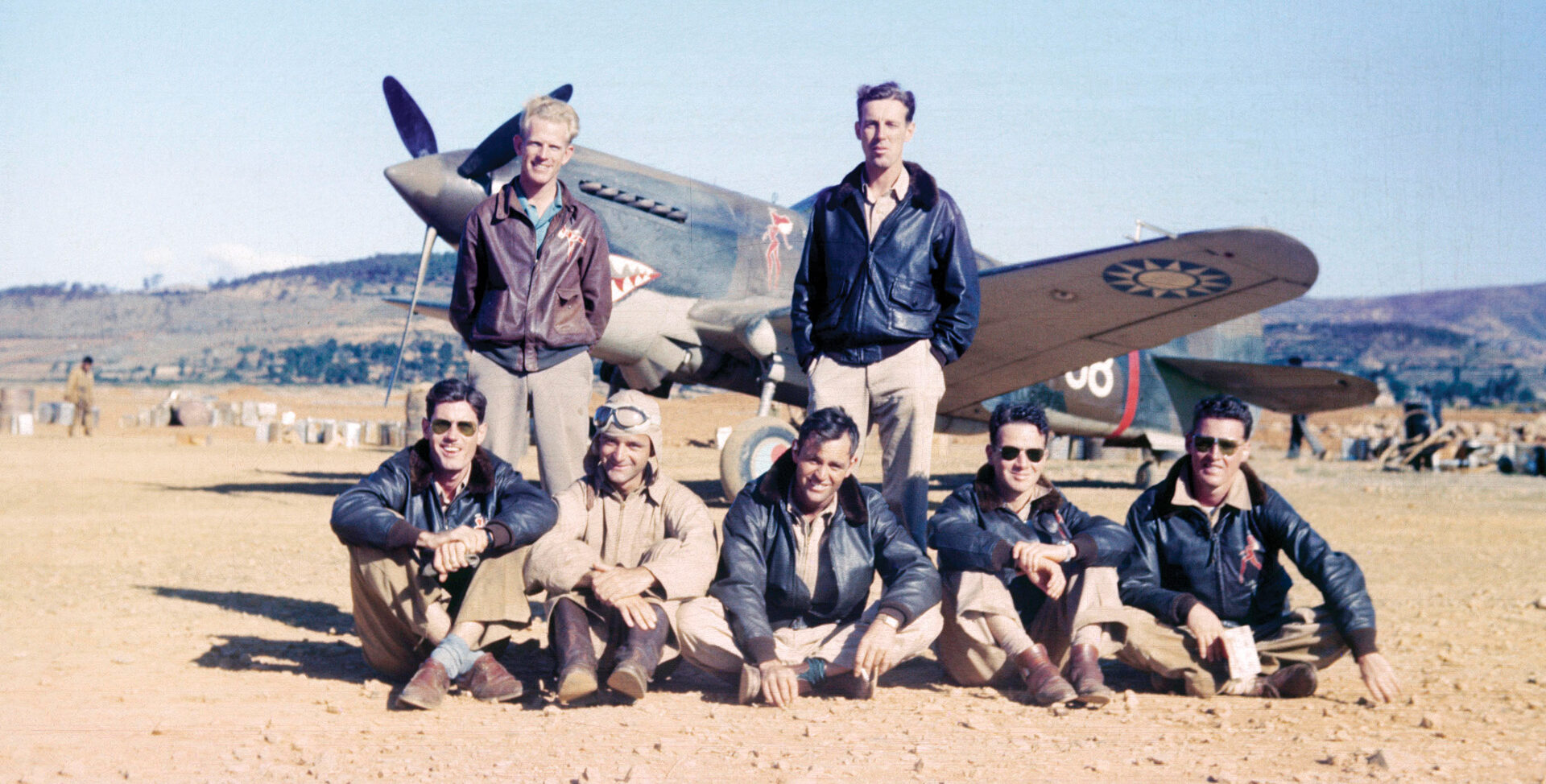 During a refueling stop at an airfield in Yunnan-Yi, China, Erik Shilling stands at left, next to Arvid Olsen, pilot of plane sitting in the background. Seated, left to right, are other members of the AVG: R.T. Smith, Ken Jernstedt, Bob Prescott, Link Laughlin, and Bill Reed.