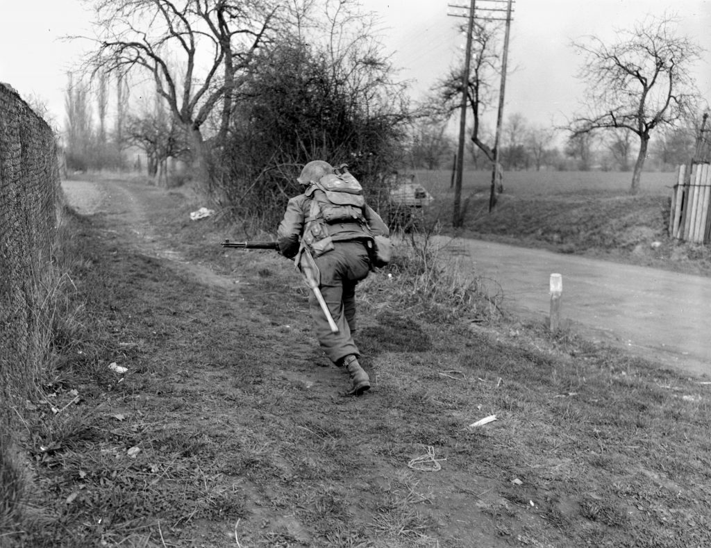 Crouching behind a stone wall for protection, an American infantryman advances along a dirt path at Aschaffenberg. The rear of the tank this soldier is accompanying may be seen on the road to the right.