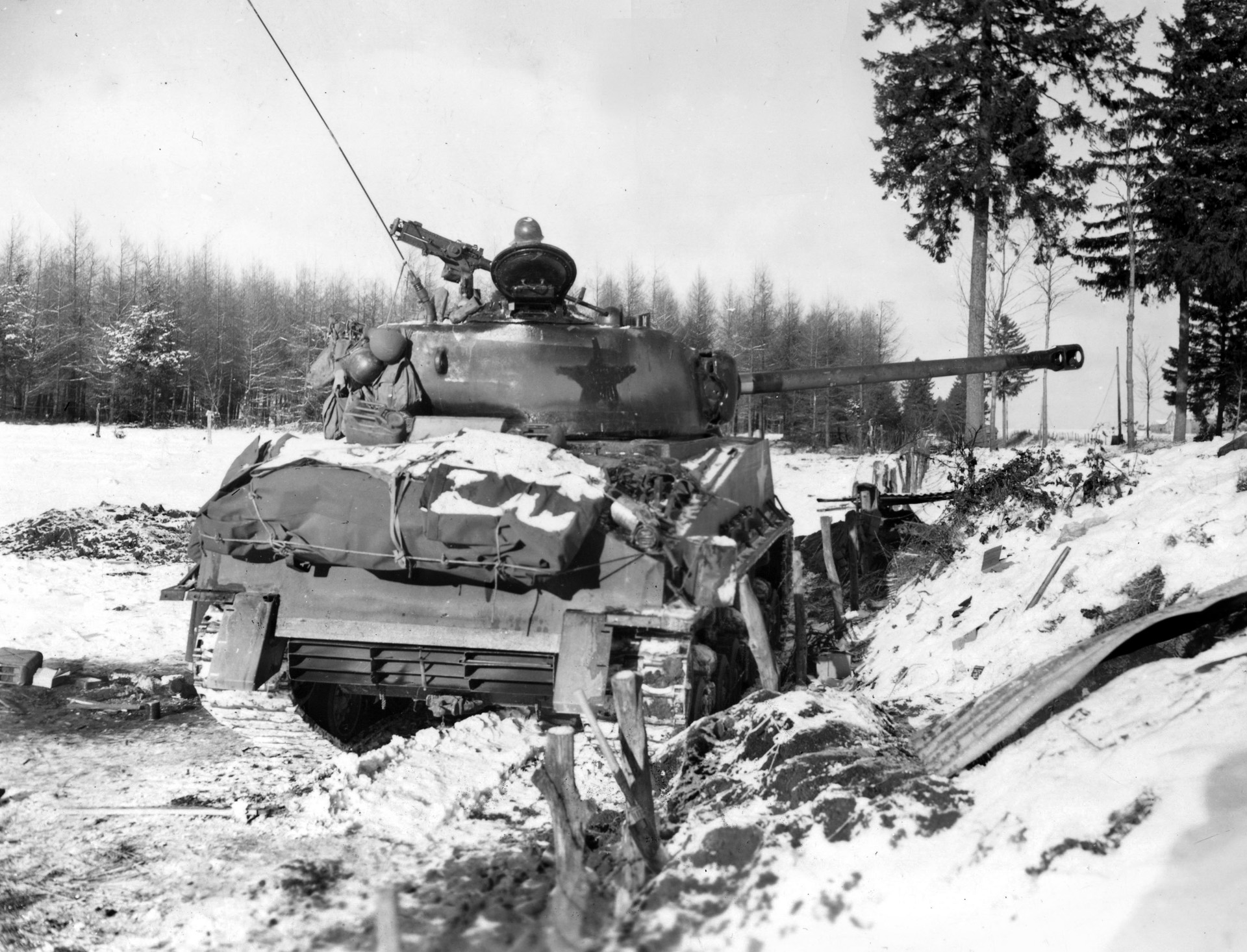 An M4A3E8 of 4th Armored Division takes cover along a sunken road while covering the H-4 highway outside Bastogne with its 76mm gun. This updated version of the Sherman has wider tracks for better performance in snow and mud; note the star has been painted over so German gunners cannot use it as an aiming point.