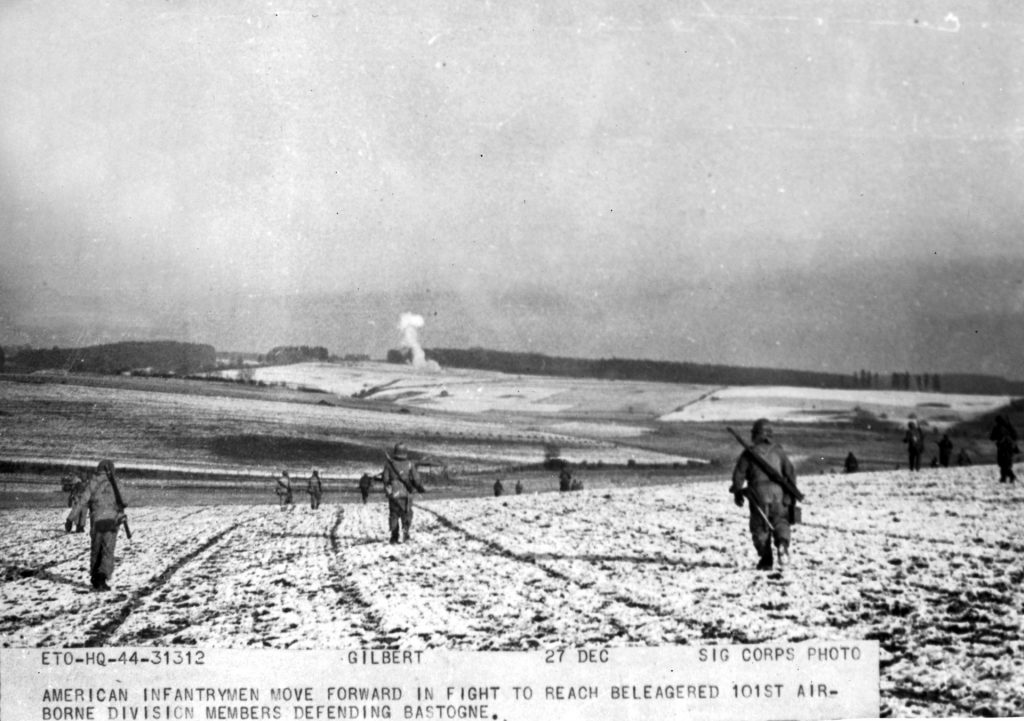 Soldiers of the 10th Armored Infantry cross a snowy field during the advance on Bastogne. A column of smoke reveals an air strike or artillery fire against the defending Germans. 