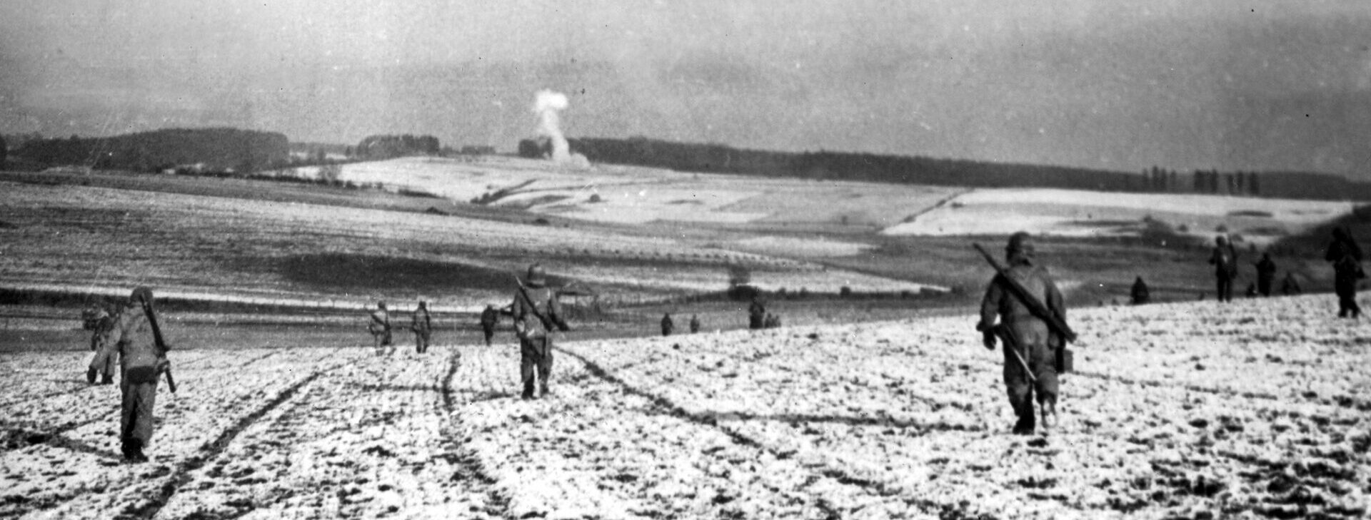 Soldiers of the 10th Armored Infantry cross a snowy field during the advance on Bastogne. A column of smoke reveals an air strike or artillery fire against the defending Germans. 