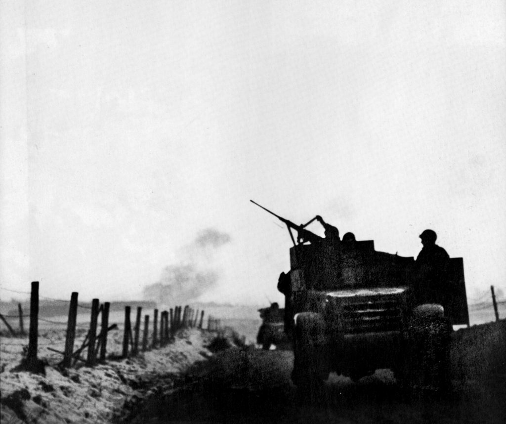 As artillery pounds a German position in the distance, a gunner on a 4th Armored Division halftrack lifts the feed tray cover on his .50-caliber machine gun to reload it. German troops feared the powerful .50 as they had nothing like it. 