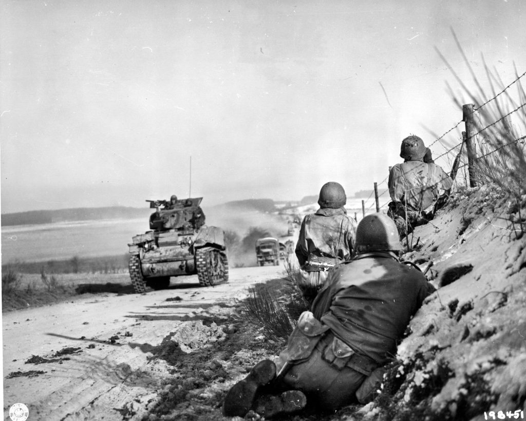 An M5A1 light tank from D Company, 37th Tank Battalion leads a convoy of trucks into Bastogne on December 27. Infantrymen guard the flanks of the corridor from German attempts to reestablish the encirclement of the town.  