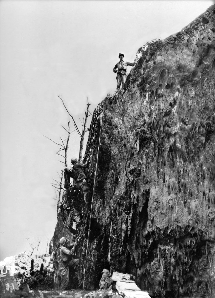 Medal of Honor recipient Corporal Desmond Doss stands on the precipice of Hacksaw Ridge as other soldiers prepare to climb to the top. Doss, a conscientious objector, served as a medic rather than carrying a rifle. While under terrific Japanese fire, he rescued dozens of wounded men during the fighting and was severely wounded himself. A feature film detailing his exploits and the battle for Hacksaw Ridge was released in 2016.