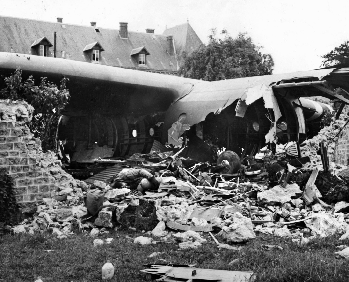 The body of a dead American soldier lies on the ground in Normandy. The soldier was apparently killed in the crash landing of the glider behind him when the aircraft careened into a stone wall.