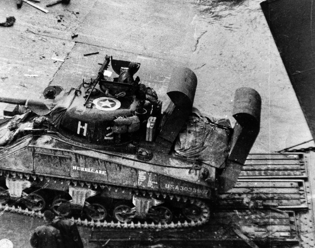 Exiting a landing craft directly onto the beach in Normandy, an American M4 Sherman medium tank prepares to advance toward the front lines. Eighteen Shermans of the 746th Tank Battalion supported Colonel Raff as he attempted to clear a landing zone for a glider-borne landing on D-Day.