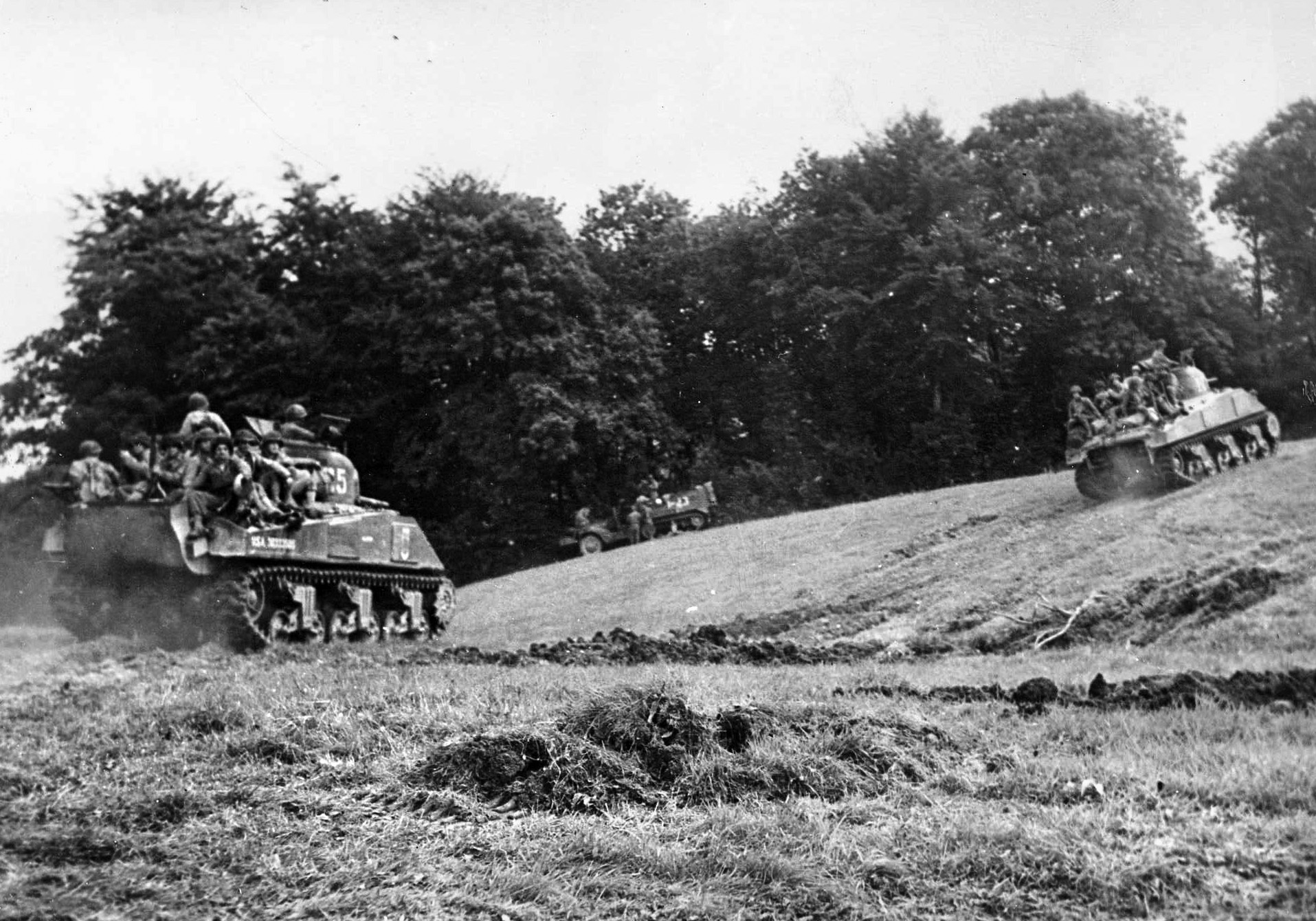 Sherman tanks negotiate a slope in the Norman countryside as American troops hitch a ride toward the front. Coming ashore with the Shermans of the 746th Tank Battalion, the men of the 401st Glider Infantry Regiment rode toward Ste-Mère-Église on the backs of the tanks.
