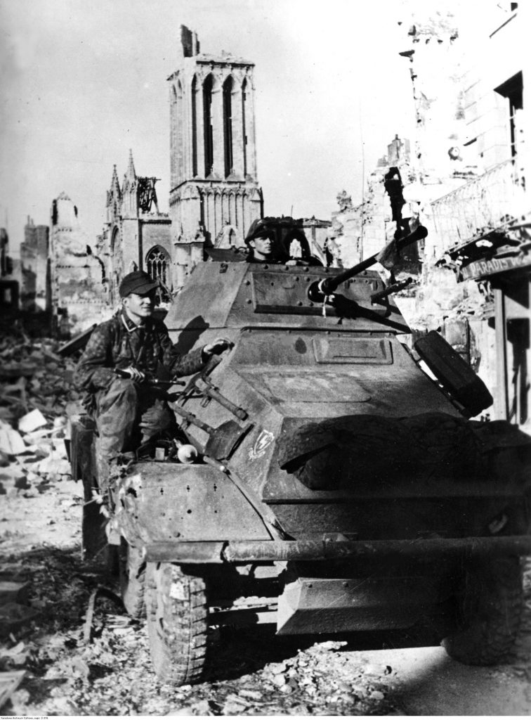 Soldiers of the 12th SS "Hitlerjugend" Armored Division ride an SdKfz 231 armored car through the devastated streets of Caen. In the background is the destroyed Church of St. Peter. 