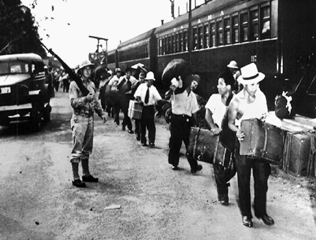 Rounded up in the Panama Canal Zone, these Japanese Latin Americans carry a few belongings and board a train while under guard. They are bound for an internment camp in the United States.