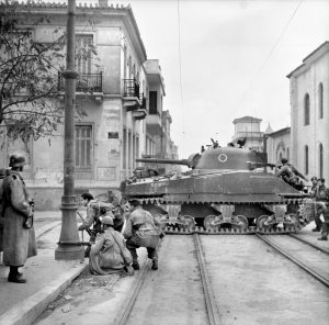 Its turret facing to the rear and its 75mm gun ready to fire, an M4 Sherman medium tank of the 5th (Scots) Parachute Battalion supports British soldiers of the 2nd Parachute Brigade during fighting in the streets of Athens against Communist ELAS militia on December 18, 1944. Two Greek soldiers, one wearing a German helmet, assist the British troops in the operation.