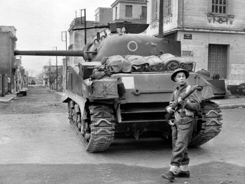 A British M4 Sherman tank sits in an intersection while soldiers remain vigilant against Communist snipers. A second British tank, covering another road crossing, is visible in the distance.