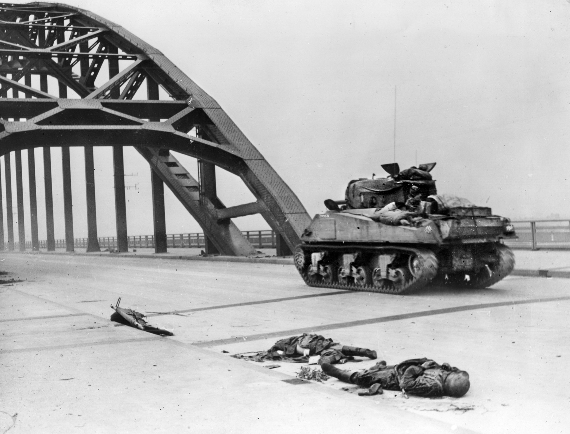 A British M4 Sherman medium tank lumbers past the bodies of two dead German soldiers, killed while defending the road bridge across the Waal River at Nijmegen. 