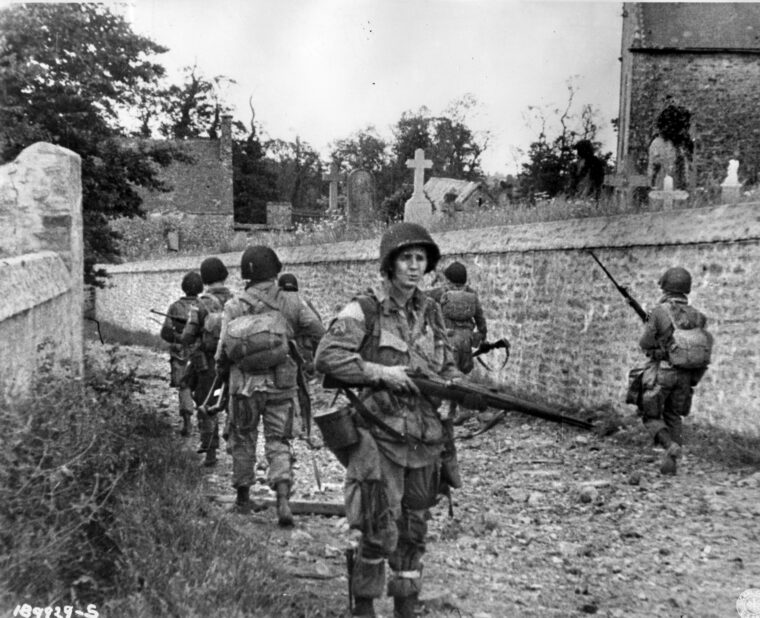 American paratroopers proceed along a dirt road through a churchyard in Normandy. In the predawn hours of June 6, 1944, the 82nd and 101st Airborne Divisions parachuted into Nazi-occupied France to seize key objectives. The focus for the 505th Parachute Infantry Regiment, 82nd Airborne was the bridge across the Merderet River at La Fiere.