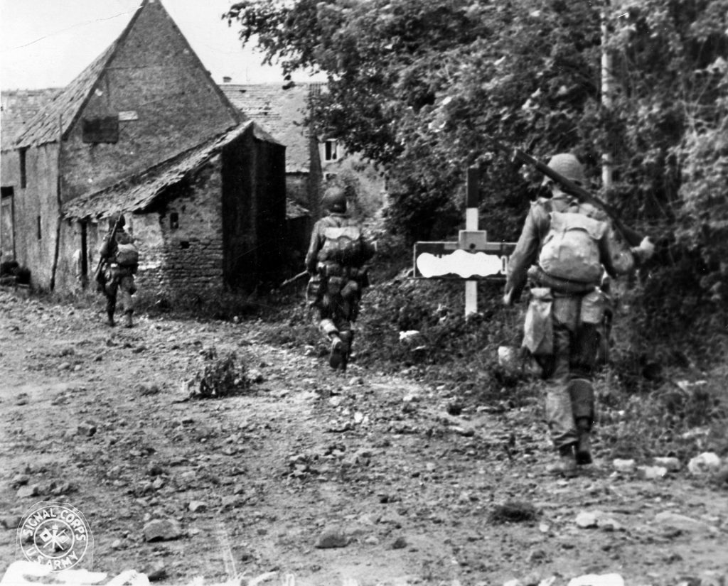 Shortly after hurtling to earth in Normandy, American paratroopers proceed down a country lane. Considerable night training, map study, and orienteering paid off when Lieutenant John Dolan’s men of A Company, 505th Parachute Infantry Regiment were able to get oriented in darkness and move south along a similar lane with a massive hedgerow providing some cover for their approach to La Fiere. Note that a censor has marked out the information on the road sign in the foreground.