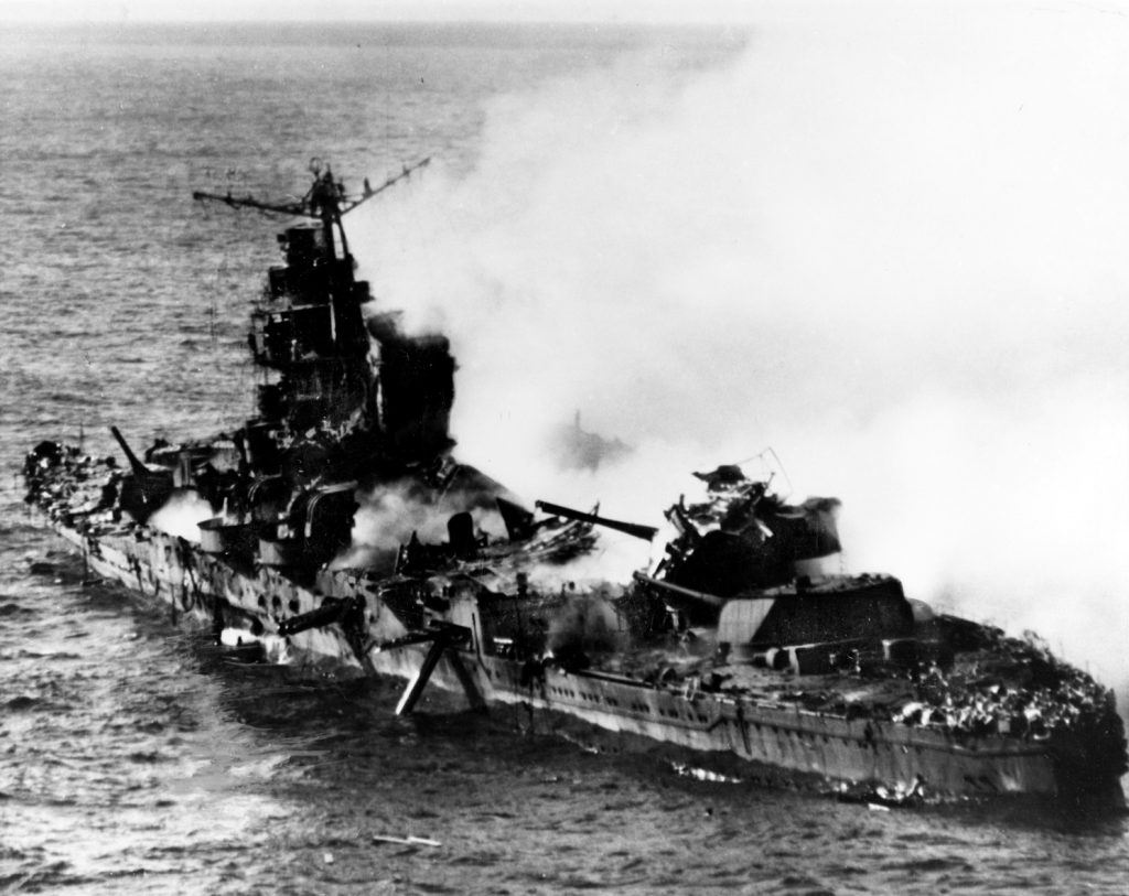The Japanese heavy cruiser Mikuma, a blackened hulk following air attacks by dive bombers from the aircraft carriers Hornet and Enterprise, is pictured on June 6, 1942, shortly before sinking during the final hours of the Battle of Midway. The roof of one of the cruiser’s eight-inch gun turrets has been blown completely off, while the barrels of the main batteries point in odd directions. The emblem of the rising sun is emblazoned atop the forward gun turret, while the superstructure is a mass of twisted steel.