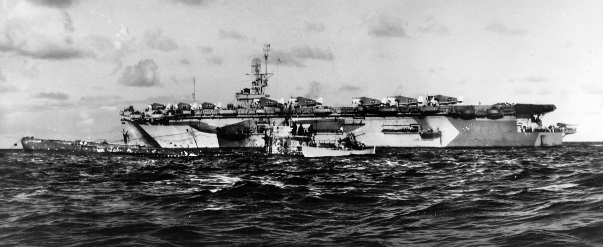 After the tow line has been successfully rigged and U-505 secured from sinking on June 4, 1944, this photo was taken depicting the captured German submarine and the escort carrier USS Guadalcanal. A small boat that carried the boarding party to U-505 remains close to the submarine.