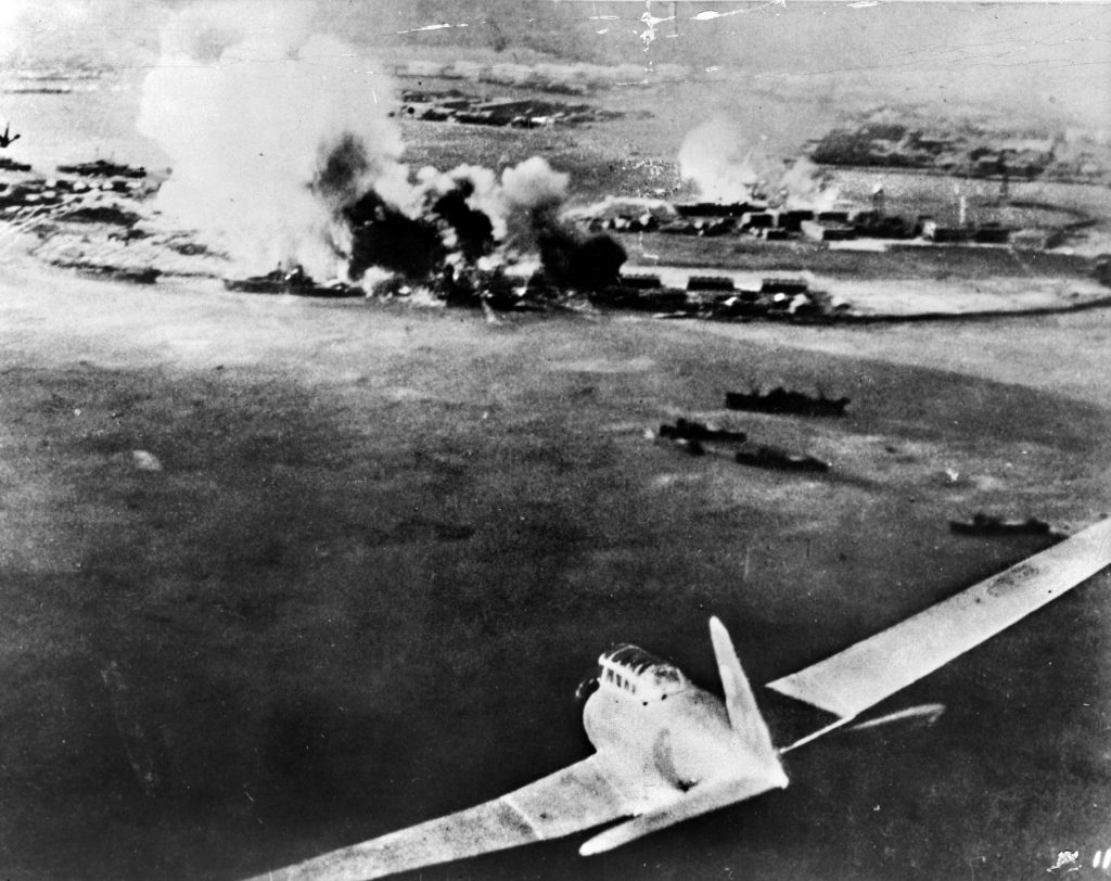 This photograph was taken by a Japanese naval aviator during the attack on Pearl Harbor. A Japanese bomber dives ahead of the plane the photographer is flying, and damage from earlier strikes is visible in the distance as heavy smoke pours from stricken facilities on the island of Oahu.