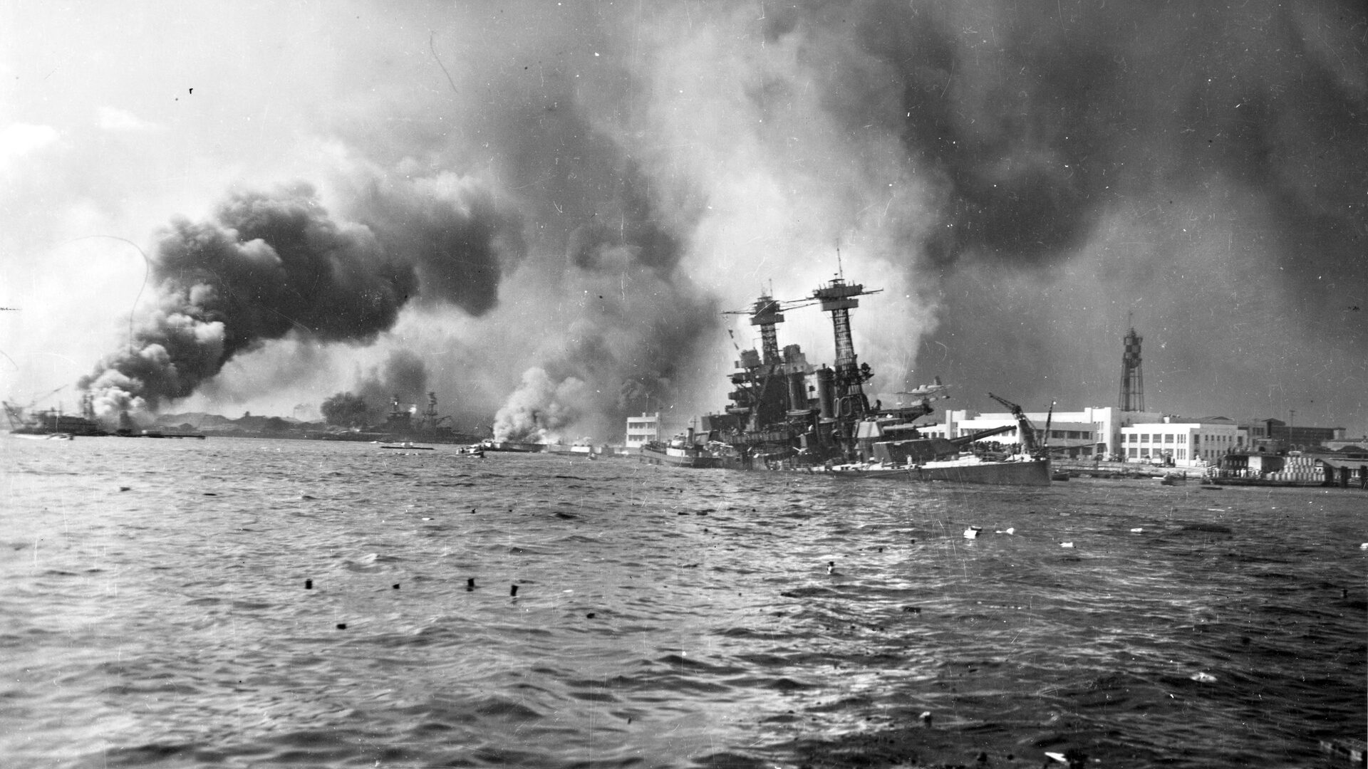 California, down by the bow, slowly sinks at its mooring adjacent to Ford Island. In the distance, the destroyer USS Shaw has exploded and burns in dry dock at far left, while the battleship Nevada, the only capital ship of the Pacific Fleet to get underway during the attack, lies beached after sustaining severe damage. 