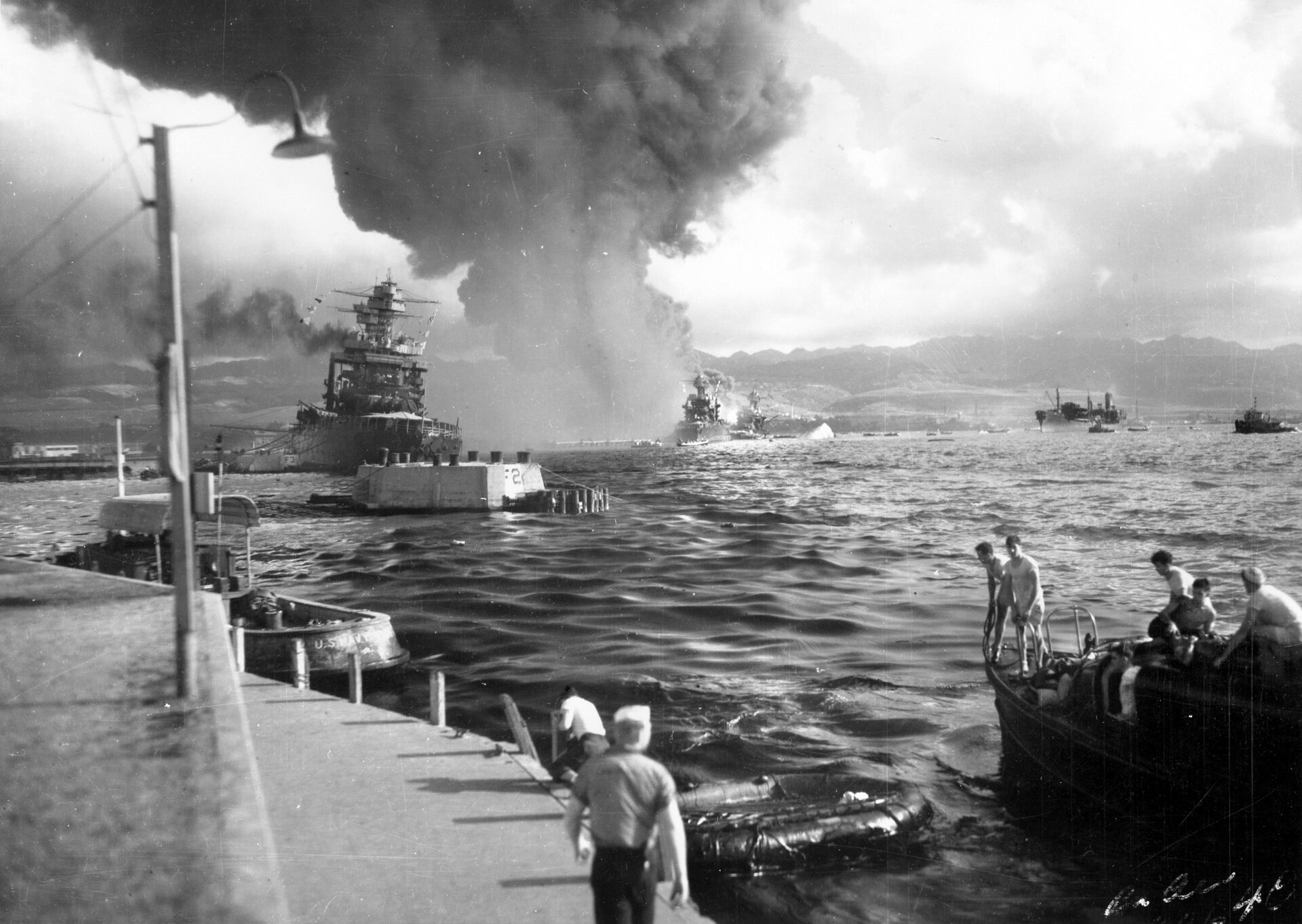 California lies listing at left in this photo after taking two torpedo hits during the Pearl Harbor attack. Beyond California lies the damaged battleship Maryland with the capsized hull of the Oklahoma to the right. The fleet oiler Neosho, in the distance at right, backs away from the devastation to remain clear of leaking oil and debris. 