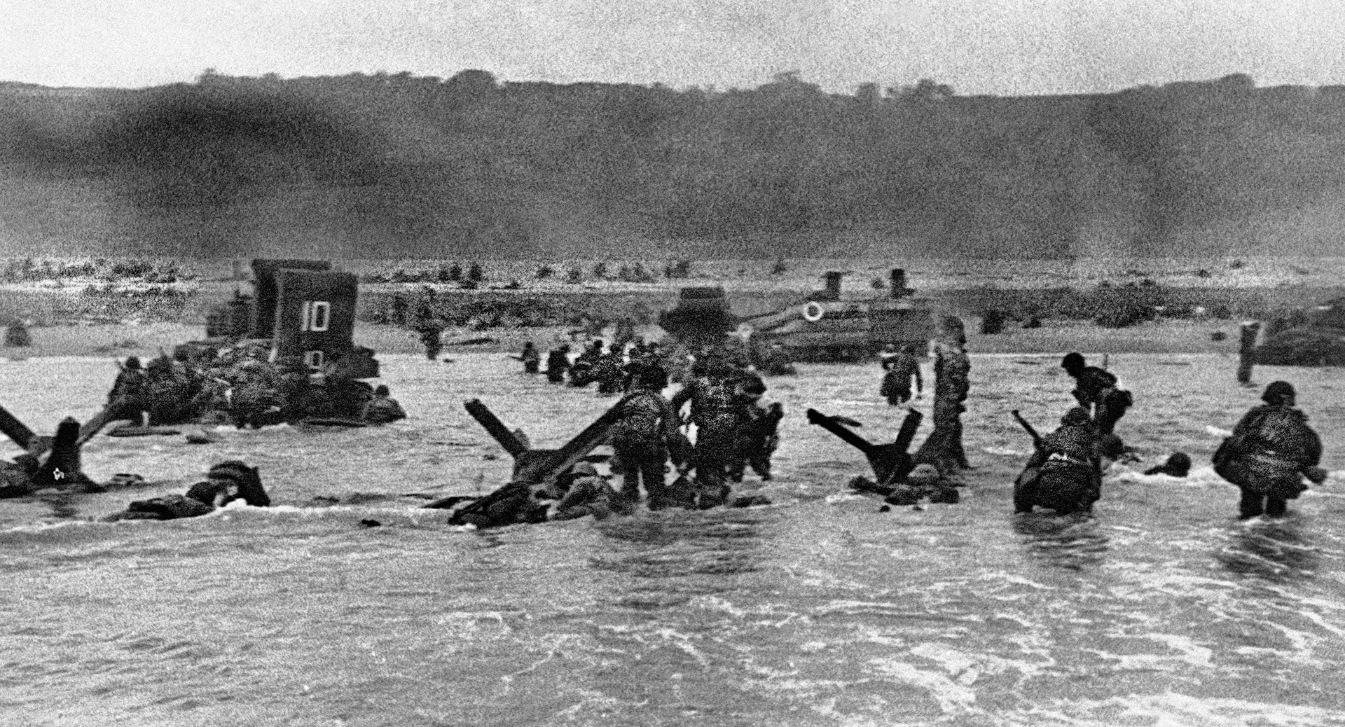  Another Robert Capa photo, taken as he departed his landing craft with the combat troops, shows men wading inland while others take shelter behind beach obstacles or three of the six DD tanks that made it to shore.