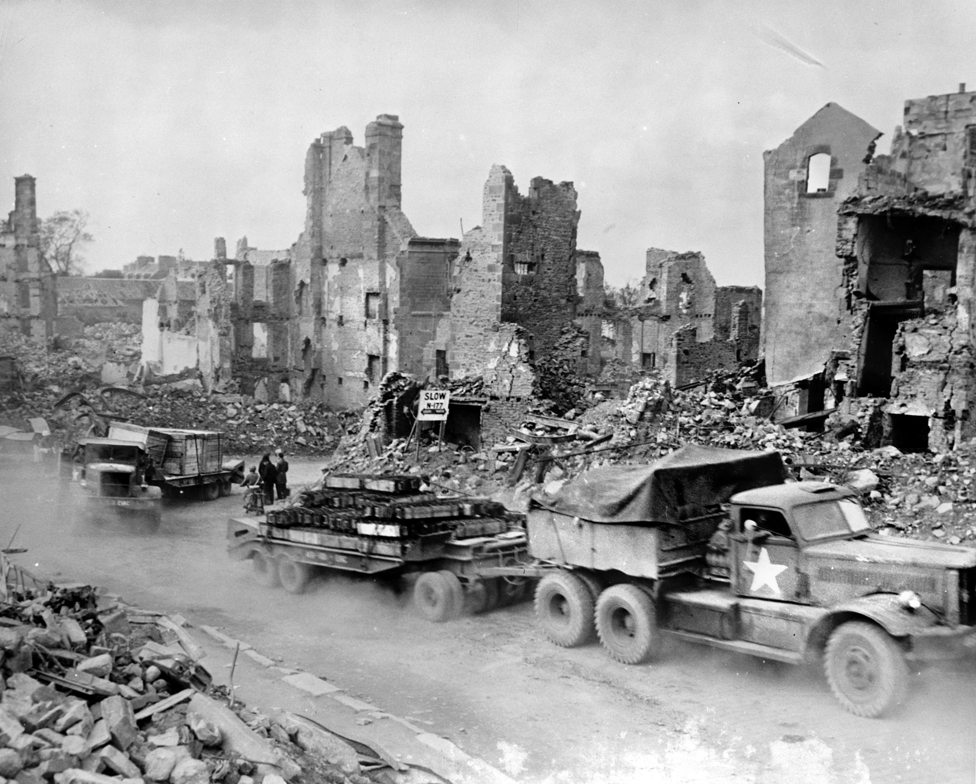 A Diamond T M20 12-ton truck and M9 trailer loaded with ammunition roars through a destroyed French town as part of a Red Ball Express convoy, France, 1944.