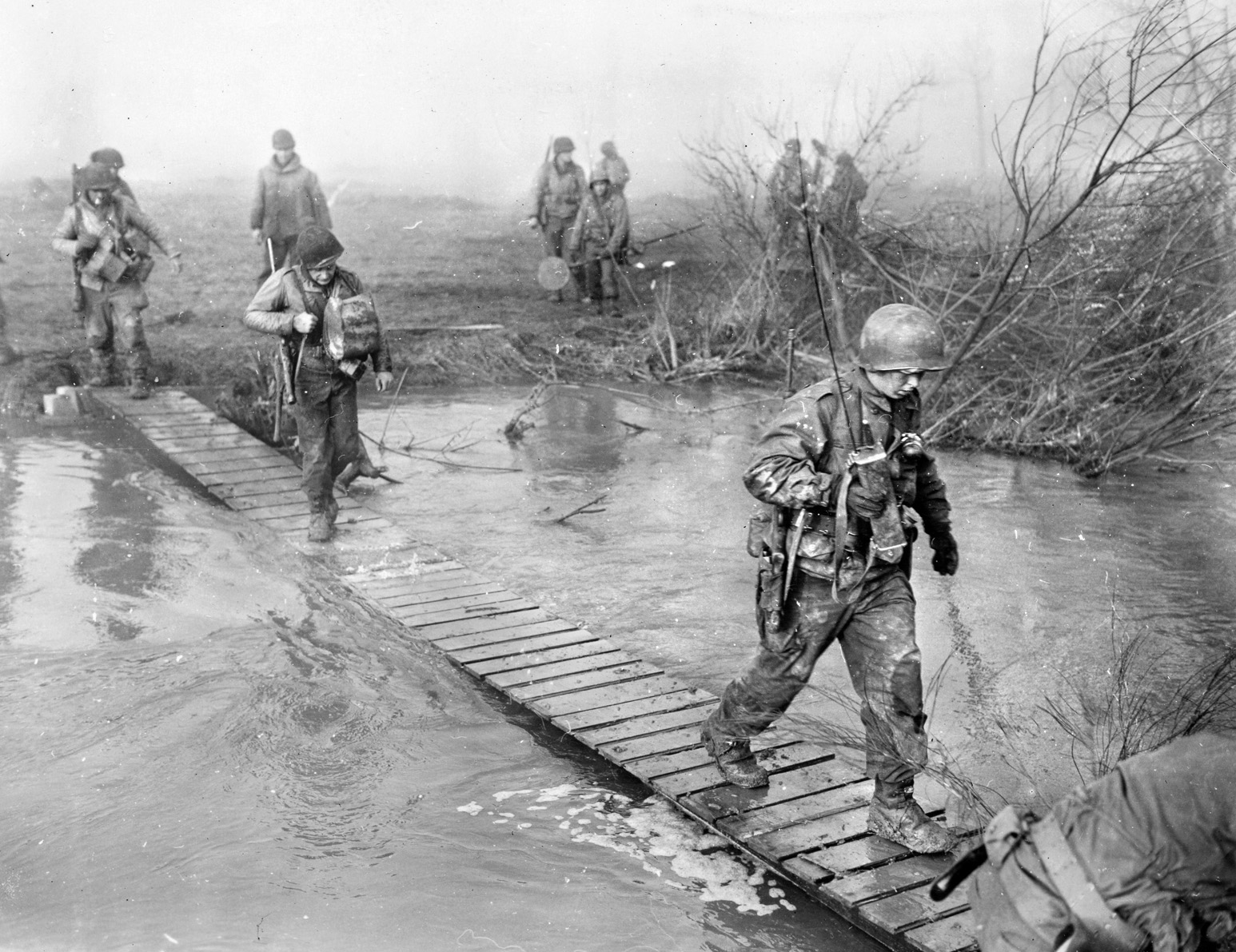 With the Germans opening floodgates and inundating the countryside, troops of the 120th Regiment, 30th Infantry Division (“Old Hickory”), cross a footbridge over the swollen Roer River, February 23, 1945.