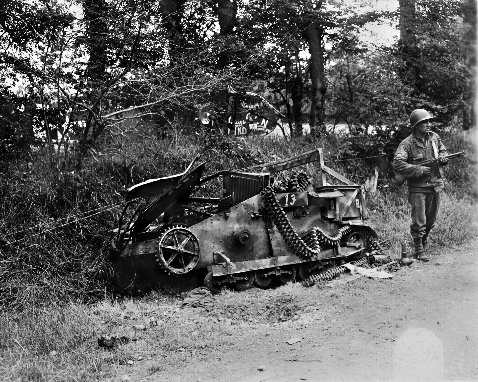 An American soldier with a Thompson sub-machine gun stands next to what appears to be a destroyed German Kettenkraftrad near Carentan. An American glider is visible through the trees.