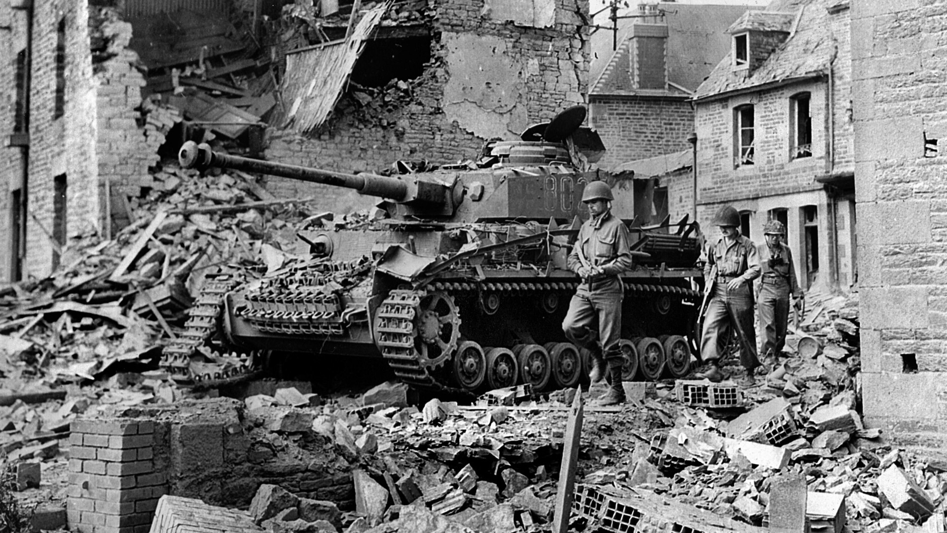 Advancing past a knocked-out Mk IV panzer, an American infantry patrol picks its way through the rubble of a Normandy village, wrecked during the Operation Cobra bombings. Cobra was launched to break through the second line of German defenses and regain the momentum lost after the initial Operation Overlord landings.