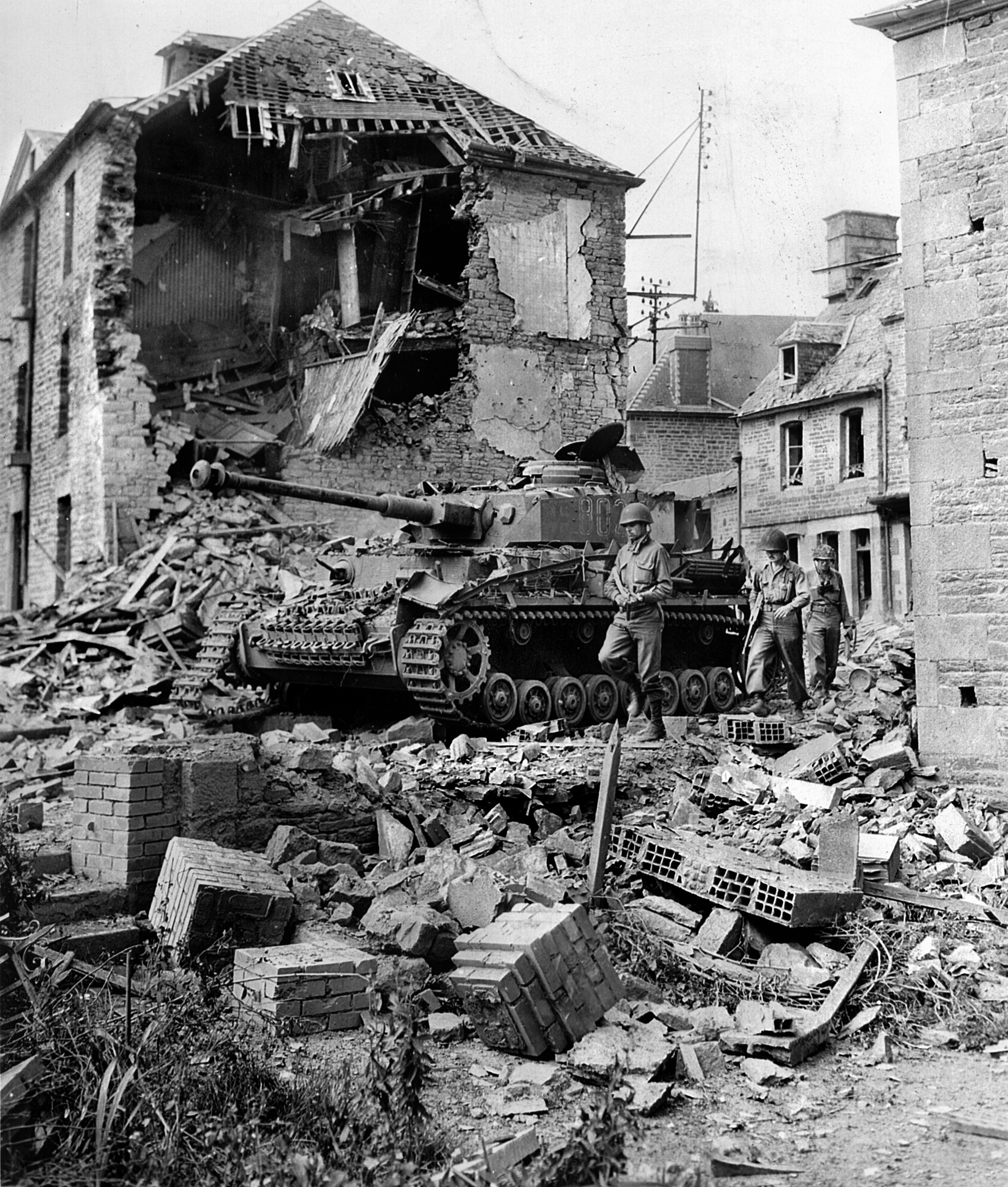 Advancing past a knocked-out  Mk IV panzer, an American infantry patrol picks its way through the rubble of a Normandy village, wrecked during the Operation Cobra bombings. Cobra was launched to break through the second line of  German defenses and regain the momentum lost after the initial Operation Overlord landings.