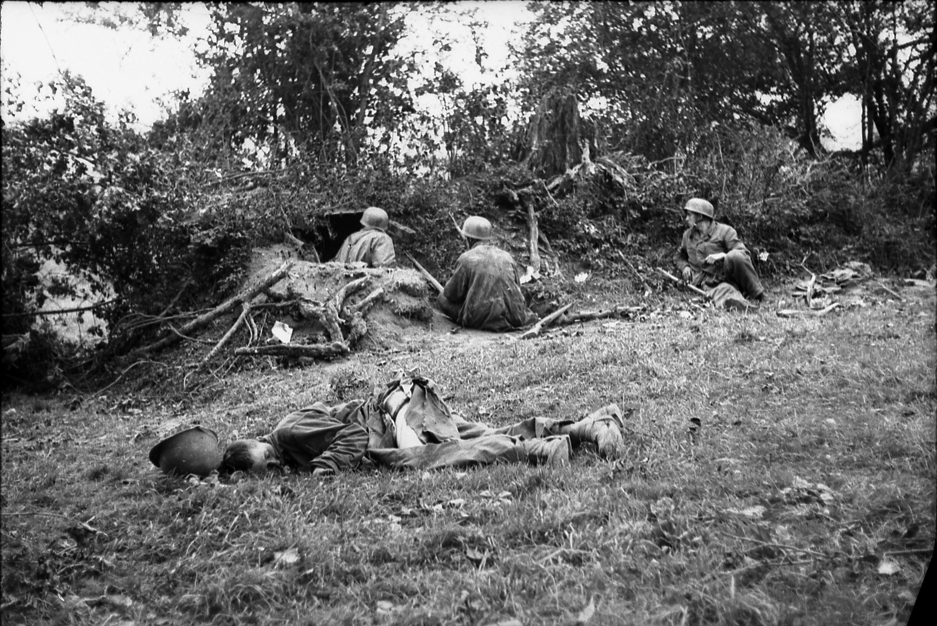 German Fallschirmjägers (paratroopers) ignore the body of a dead American and take cover behind a thick hedgerow in Germany. The hedgerows were unexpected obstacles faced by the invaders, bogged down the advance.