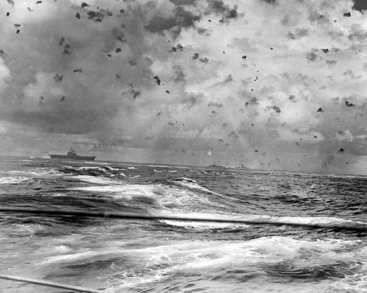 Black puffs of exploding antiaircraft shells dot the sky above Kinkaid’s TF 61 as U.S. Navy ships try to fend off swarms of attacking Japanese planes. USS Enterprise is at left with at least two enemy warplanes overhead. Ship second from right is USS South Dakota, firing her starboard 5/38 secondary battery, as marked by the bright flash amidships. 