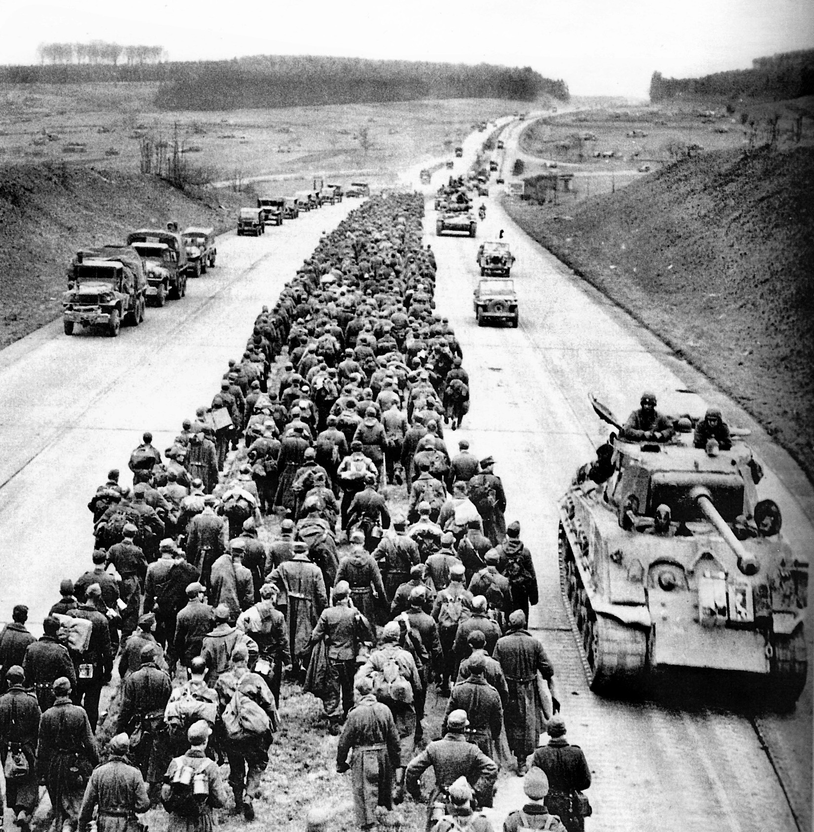 American trucks, tanks, and jeeps roll eastward along an autobahn while thousands of German troops march west down the median on their way to a POW enclosure. 
