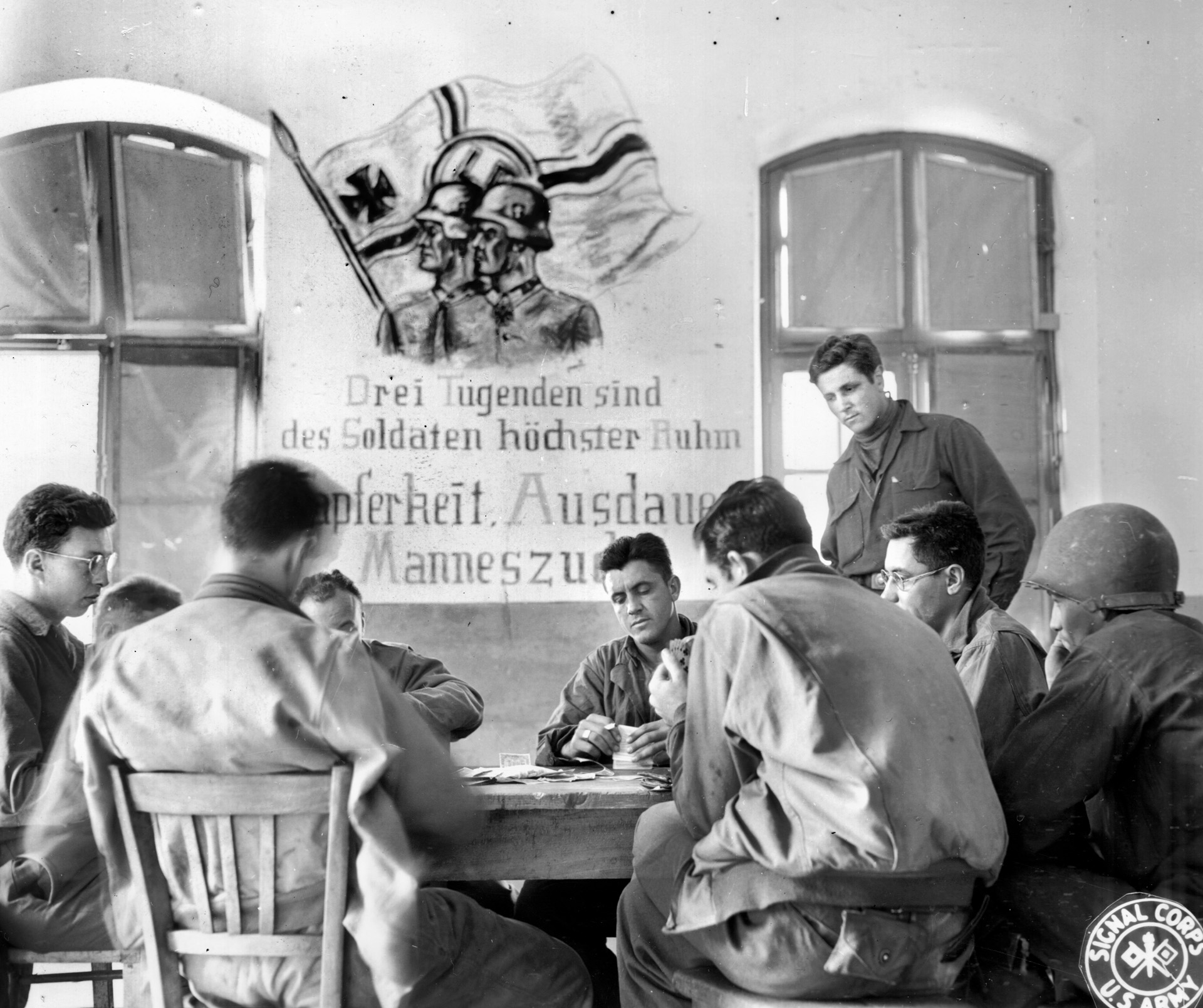 A group of American soldiers enjoys a spirited game of poker in a building in Luneville that was once occupied by German troops.  The poster on the wall, meant to inspire the German troops, reads: ‘Three virtues are the soldier’s greatest fame:  bravery, perseverance, and manhood.’ 