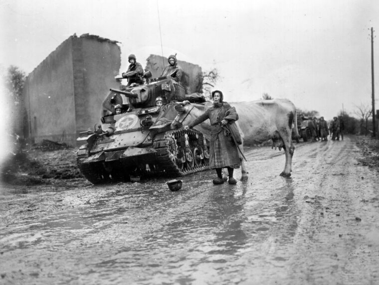 A tank crew of the 2nd Cavalry Regiment waits patiently for a French woman and her cow to cross a rain soaked road near the town of Luneville in the fall of 1944. The regiment was a component of the 4th Armored Division and played a key role in disrupting a German attempt to cut the 4th Armored off and destroy it. A derelict German helmet lies in the roadway.