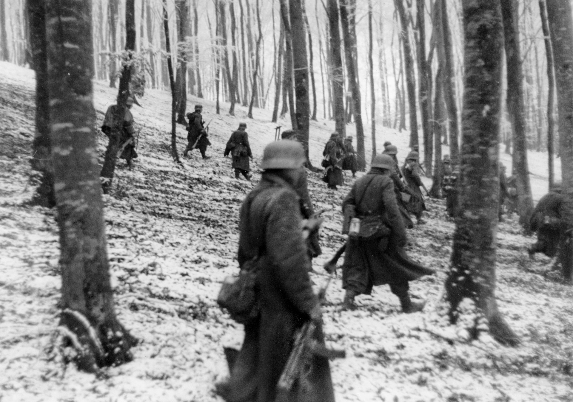 German infantrymen, their field-gray overcoats standing out against a snow-covered hillside, relocate during operations in Luxembourg in December 1944.