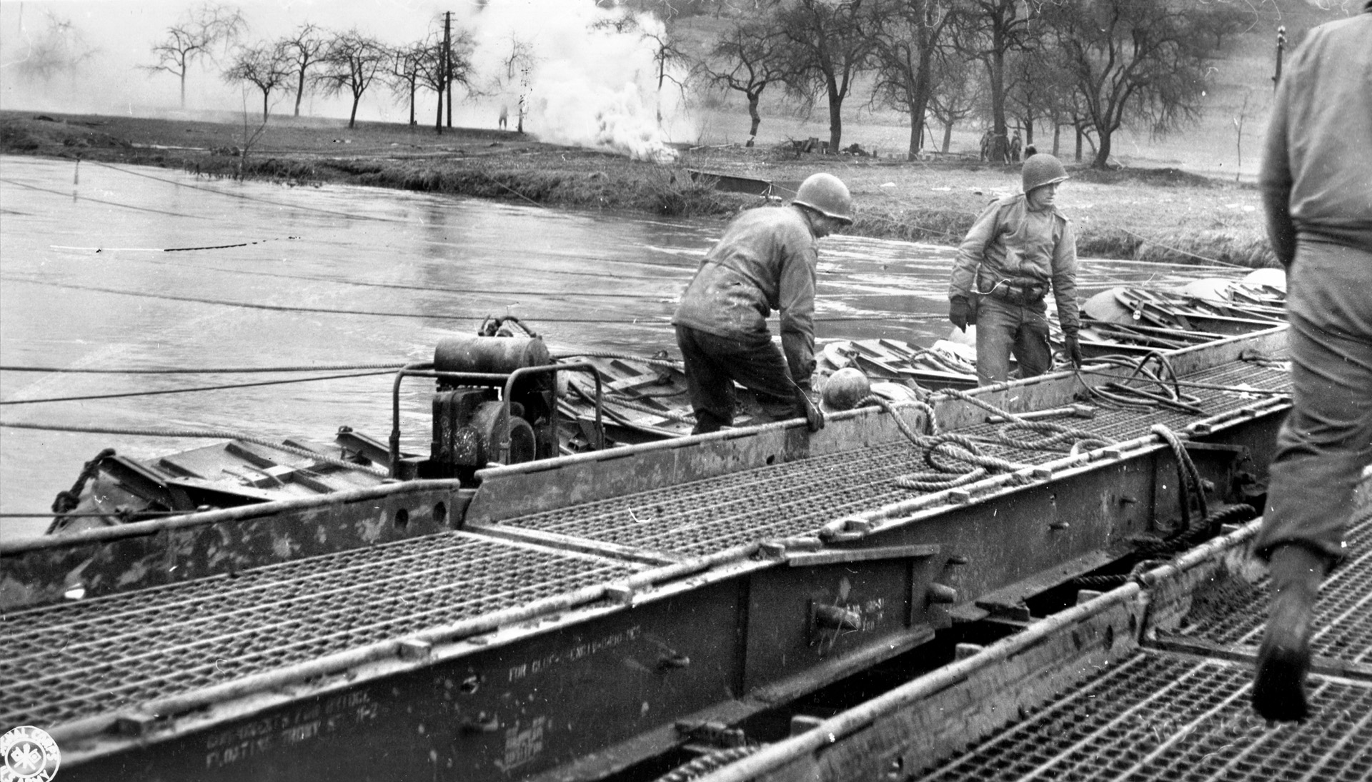 American combat engineers with the 5th Infantry Division erect a treadway bridge across the Sauer River. Smoke, intended to help protect the exposed engineers from enemy fire, billows after a shell has impacted the opposite bank. 