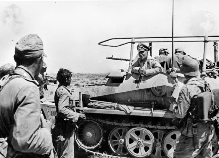 Field Marshal Erwin Rommel visits with troops of the Africa Korps while inspecting positions in the Tobruk fortress belt. Rommel is aboard his light-infantry command vehicle SdKfz. 250/3 ‘Greif.’ The vehicle is equipped with radio-communications gear; note the antenna apparatus atop the open personnel area.
