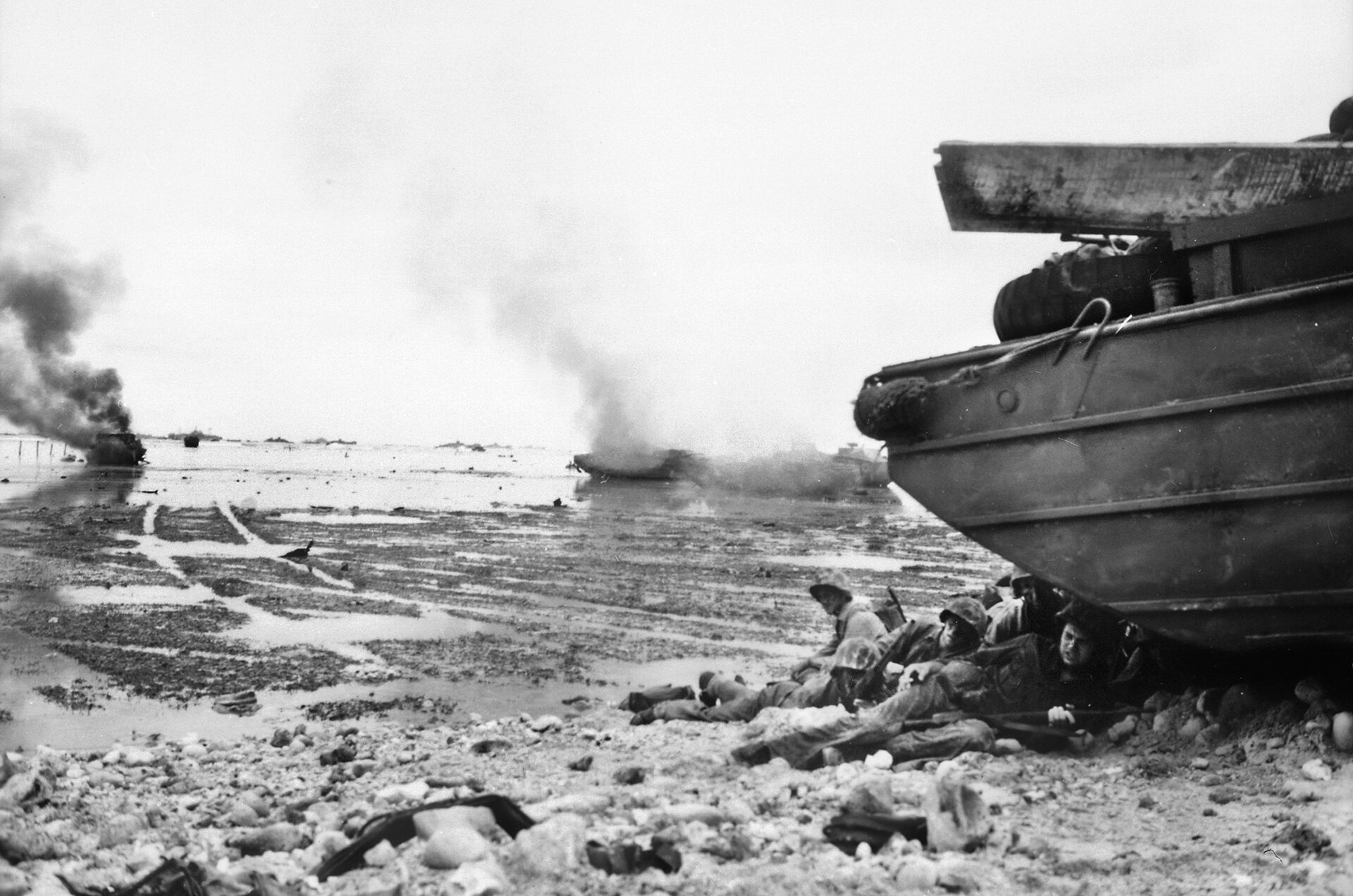 American Marines take cover from intense Japanese fire on the beach at Peleliu. These men are crouching behind an amphibious DUKW vehicle, while a landing craft burns furiously in the background after receiving a direct hit from a hidden Japanese gun on shore. 