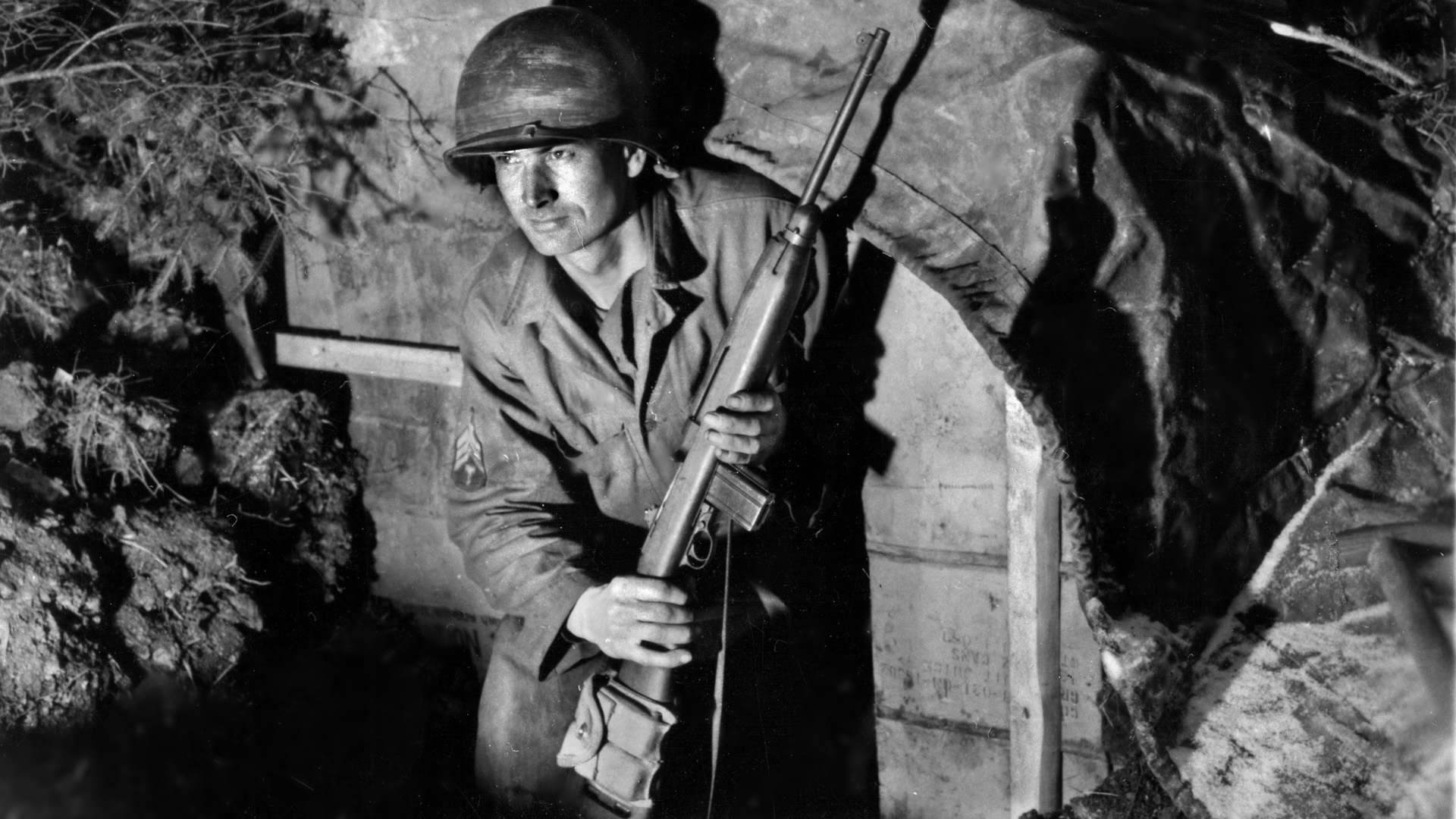 Sergeant Fred Parke of the U.S. 99th Infantry Division, armed with an M-1 carbine, peers from the dugout he occupies during the Battle of the Bulge in Belgium. The I&R Platoon of the 99th Division’s 394th Infantry Regiment held off superior German forces at the village of Lanzerath for a full day.