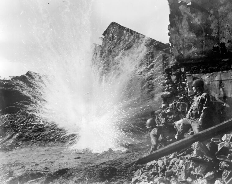 A German Nebelwerfer, also known as a Screaming Meme, explodes near a group of soldiers outside a Belgian village. During the battle for Noville, a Screaming Meemie exploded near Freeman, injuring his leg and taking him out of the campaign.