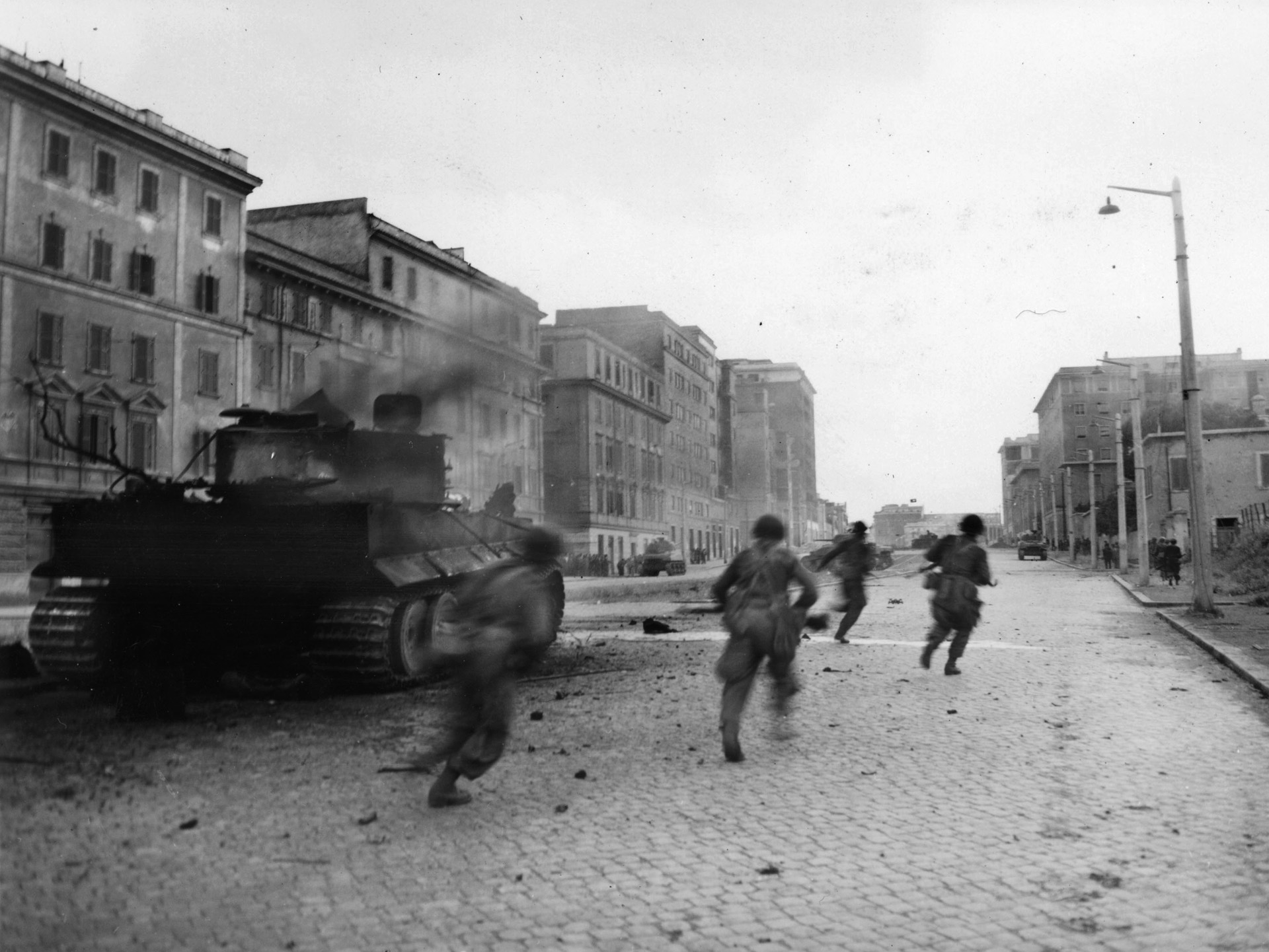 American soldiers, among the first Allied troops to enter Rome, sprint past a burning German tank to take up positions along a city street. An American tank, providing fire support as the infantrymen clear houses, is seen in the distance at right.
