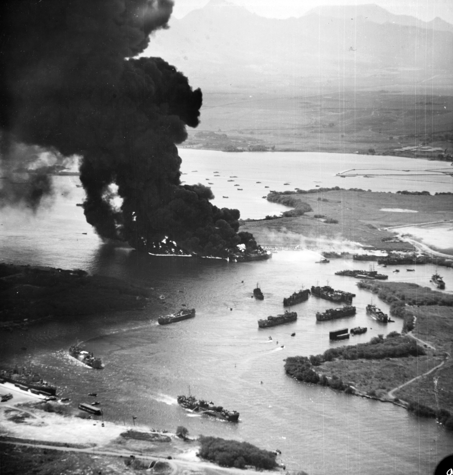 This aerial view of Pearl Harbor’s West Loch in flames clearly shows burning LSTS at Tares 8 and 9 while other LSTs attempt to rapidly maneuver away from the conflagration toward safety. Several vessels clustered to the right in this image appear to remain stationary despite the threat.