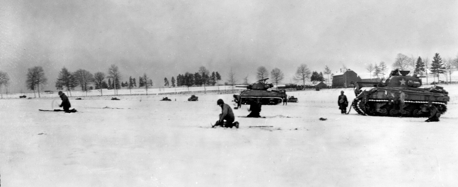 During heavy fighting against German forces in the vicinity of Bastogne, armored infantrymen of the 6th Armored Division advance cautiously across a snow-covered meadow. Sherman tanks are visible in support of the advance.