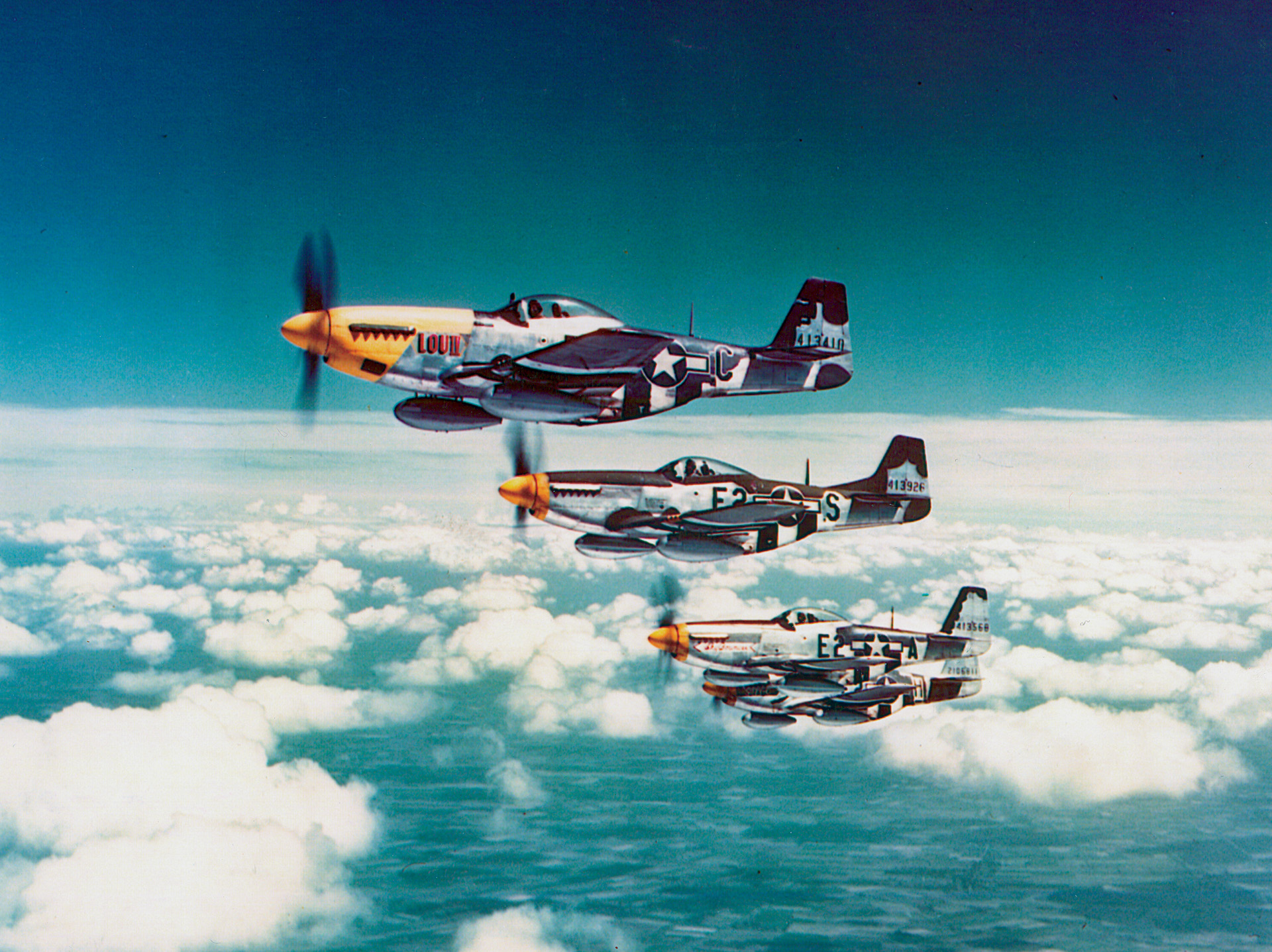 Carrying drop tanks to extend their range, a flight of P-51 Mustangs maintains formation in the skies over Europe. The P-51 became the premiere Allied long-range escort fighter of World War II, even reaching Berlin from airfields in England.
