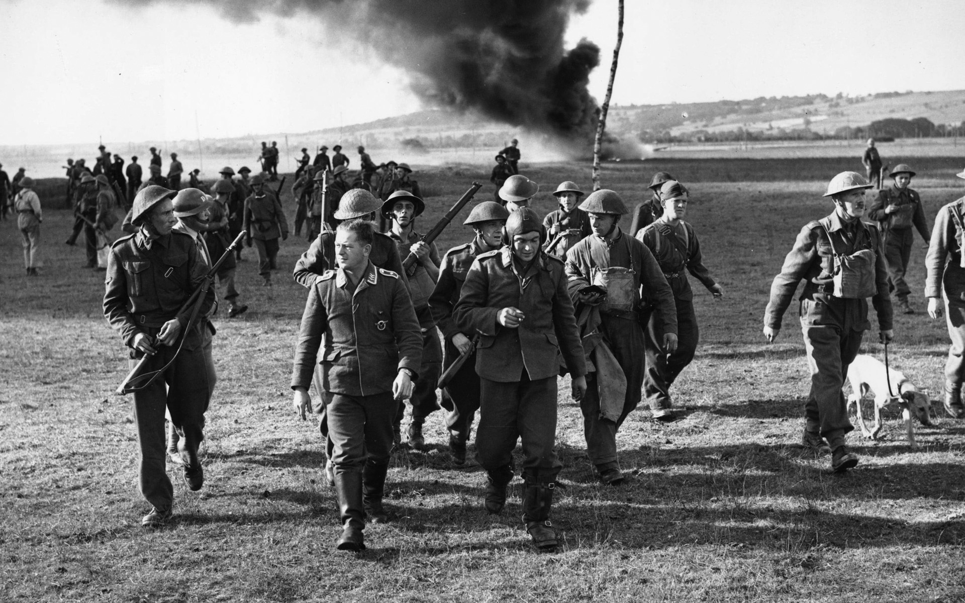 Two German airmen, who parachuted from their shot-down Heinkel He-111 bomber (burning in the background) are marched off by the Home Guard in Goodwood, Sussex.