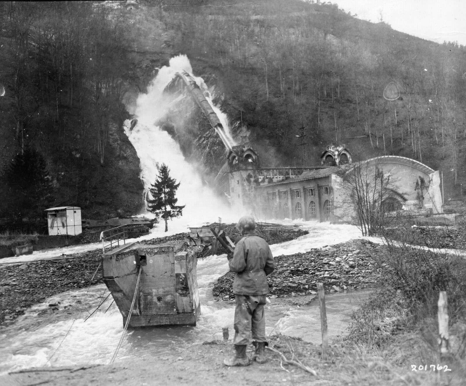 An American soldier watches torrents of water from the River Rur flow around the Heimbach hydro electric plant after the Schwammenauel Dam was breached in February 1945.